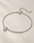 The Eden Moissanite Bracelet in polished silver tone, arranged as a full loop with a curved bar, one round clear stone, ball-bead chain, and extension clasp on a pale-greige background, reference-based AI visualization.