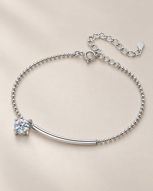 The Eden Moissanite Bracelet in polished silver tone, arranged as a full loop with a curved bar, one round clear stone, ball-bead chain, and extension clasp on a pale-greige background, reference-based AI visualization.