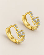 The Veyra Geometric Huggies, two warm yellow-gold-coloured geometric huggies in a low-angle diagonal resting arrangement with clear rectangular and round stones on pale greige, reference-based AI visualization