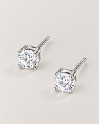 The Neva CZ Stud Earrings — silver-tone diagonal pair view with clear round stones and visible posts in a pale-greige studio; reference-based AI visualization.