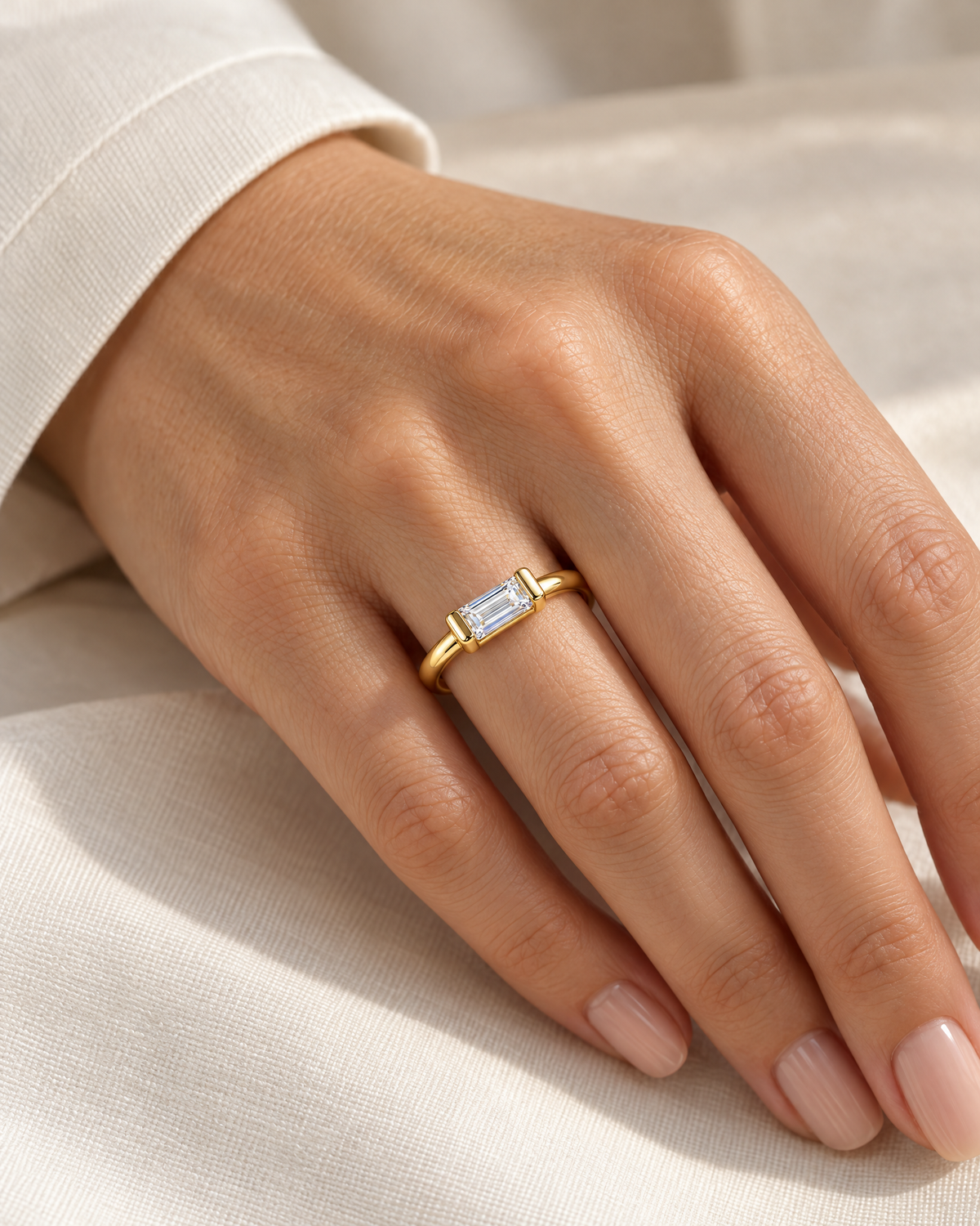 The Lyra CZ Emerald-Cut Statement Ring — gold-color clear emerald-cut ring worn on a relaxed light-warm adult hand with ivory linen; reference-based AI visualization.