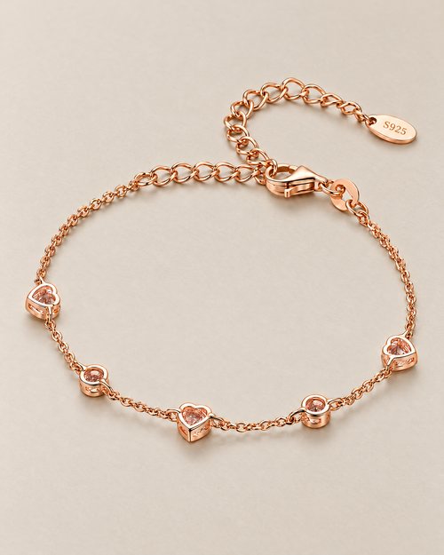 The Dahlia S925 Heart Bracelet in warm rose-gold-toned metal, shown from the open-backed side with five alternating heart and round stations in an oval on pale greige, reference-based AI visualization.