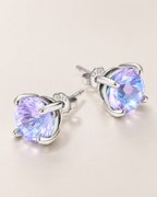 The Frida Firework CZ Stud Earrings: purple-lilac faceted four-prong stud pair with posts, scalloped butterfly backs and visible S925 marks on pale greige, reference-based AI visualization