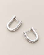 The Aria CZ Hoops — separated diagonal polished silver-colored U-shaped hoop pair with clear round stone rows on pale greige, reference-based AI visualization