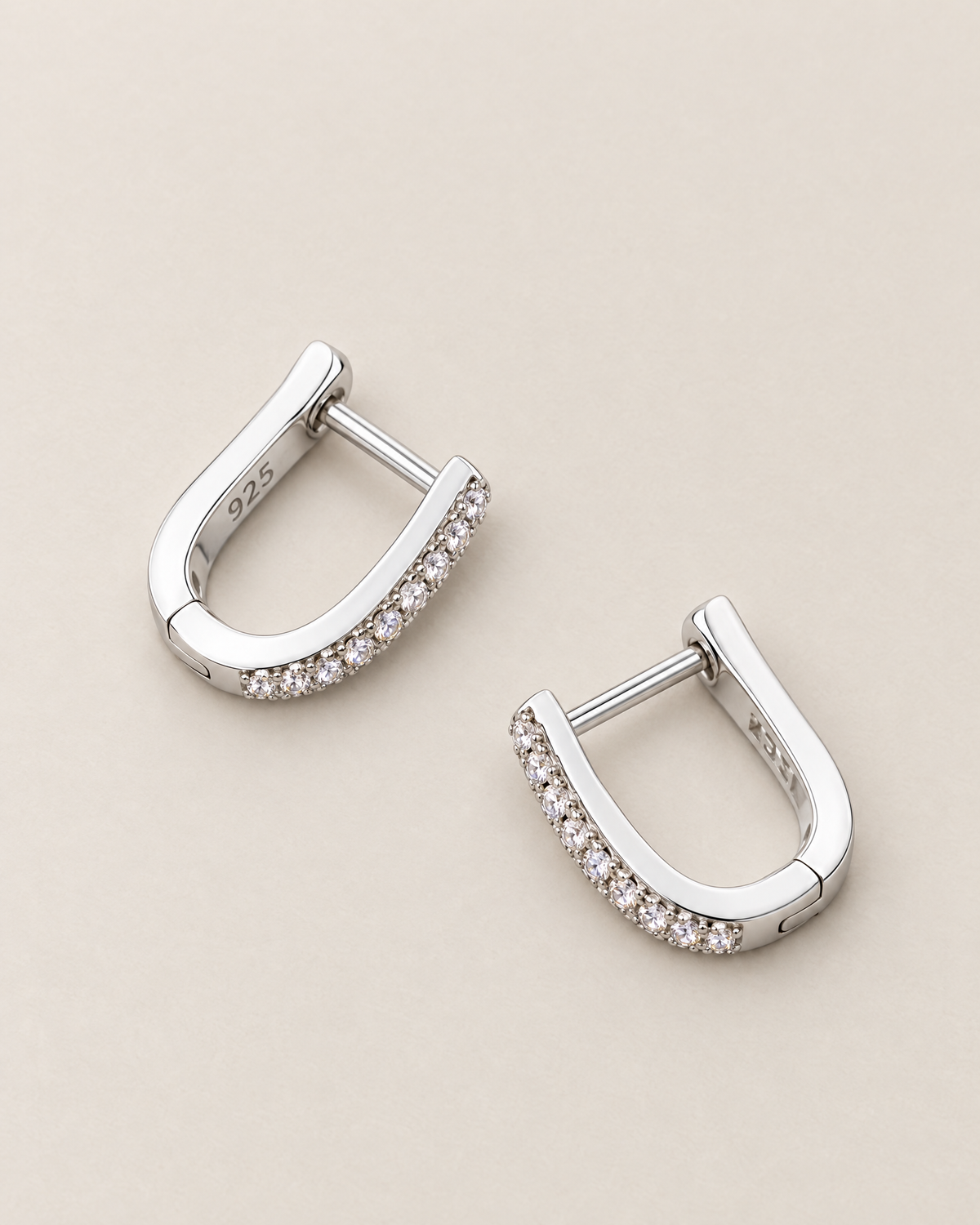 The Aria CZ Hoops — separated diagonal polished silver-colored U-shaped hoop pair with clear round stone rows on pale greige, reference-based AI visualization