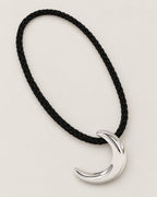 Black braided cord arranged in a diagonal oval around a polished crescent pendant on pale greige, reference-based AI visualization