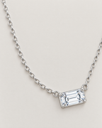 The Mara Geometric CZ Pendant — jewelry-only spacious overhead arrangement with a horizontal clear rectangular stone and alternating-link chain on pale warm greige; reference-based AI visualization