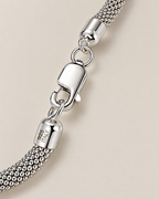 The Liva Mesh Chain Bracelet clasp detail in silver-tone metal with woven mesh, oval connector rings, and visible 925 ITALY end-cap marking on warm pale greige; reference-based AI visualization.