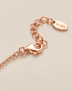 The Dahlia S925 Heart Bracelet in warm rose-gold-toned metal, close on its lobster clasp, extension links, and marked oval end tag on pale greige, reference-based AI visualization.