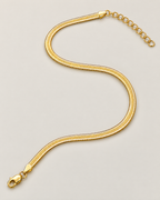 Gold-tone flat herringbone necklace in a flowing open S-shaped lay with clasp and extender at opposite ends on a pale greige studio ground; reference-based AI visualization