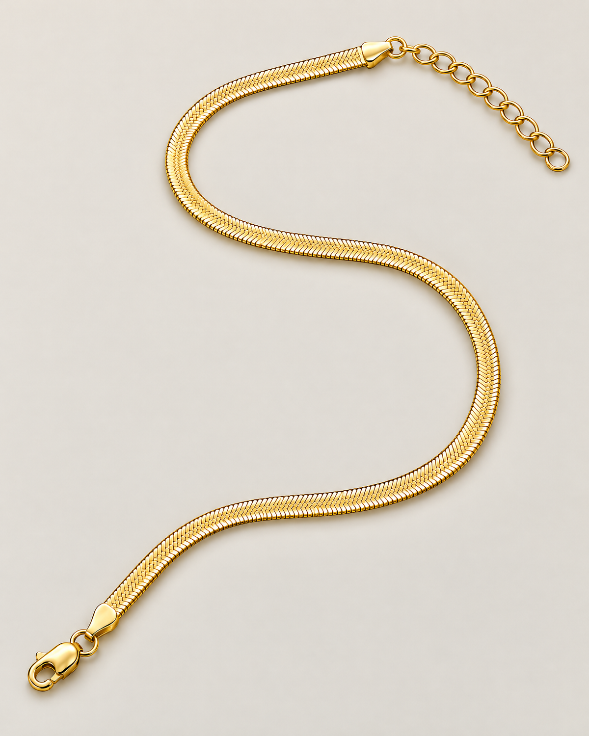 Gold-tone flat herringbone necklace in a flowing open S-shaped lay with clasp and extender at opposite ends on a pale greige studio ground; reference-based AI visualization