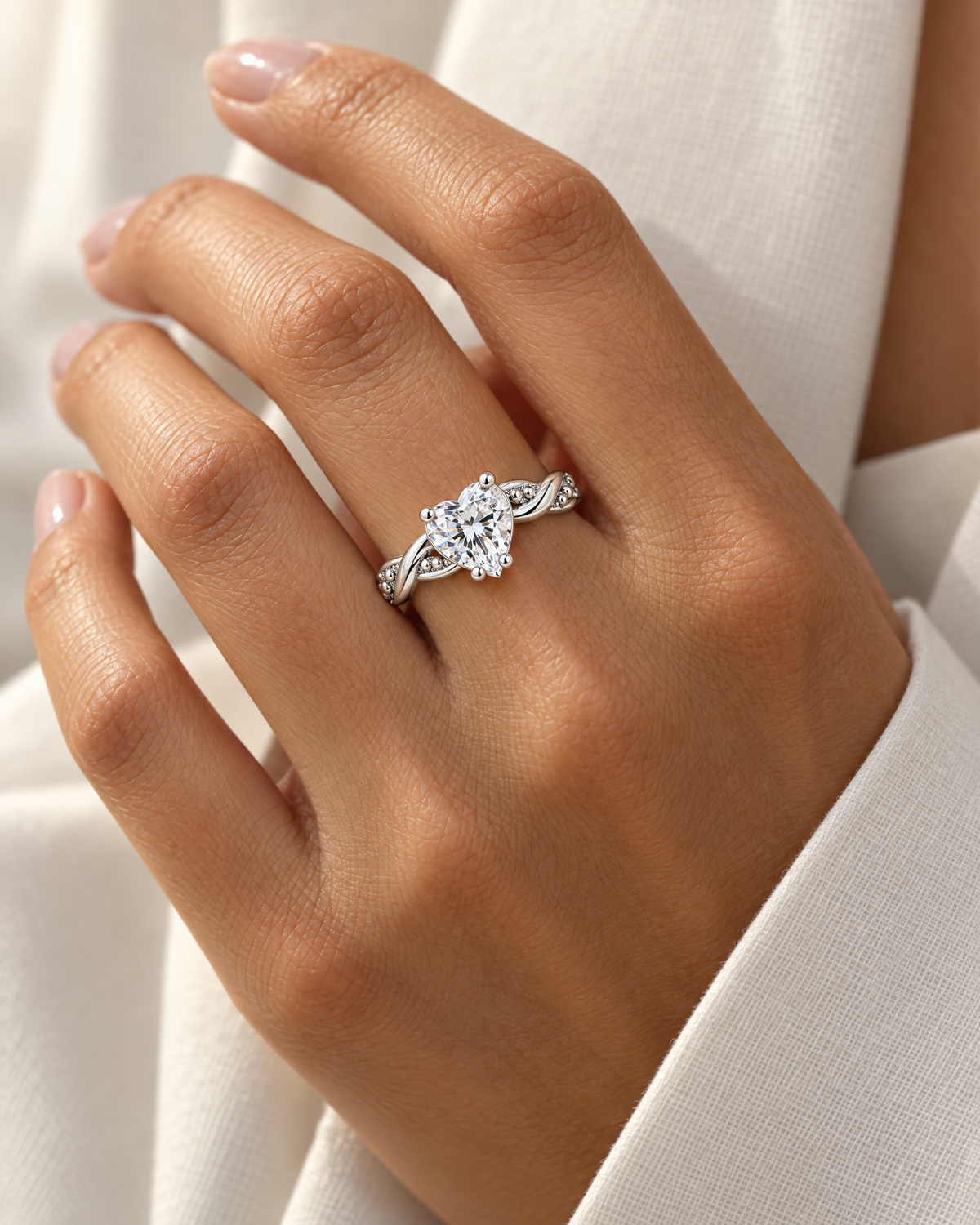 The Alva Heart-Stone Ring, observed polished silver-tone with a clear heart-shaped center stone and twisted beaded shoulders, worn naturally on a medium-warm adult hand with an ivory linen cuff — reference-based AI visualization.
